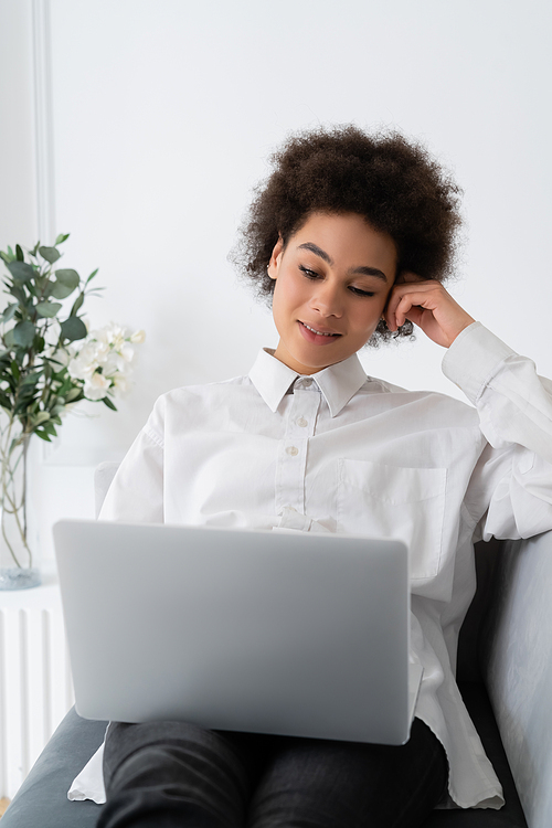 cheerful african american woman using laptop while sitting on grey velvet sofa