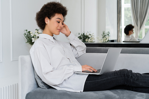 african american woman using laptop while sitting on grey velvet sofa and working from home