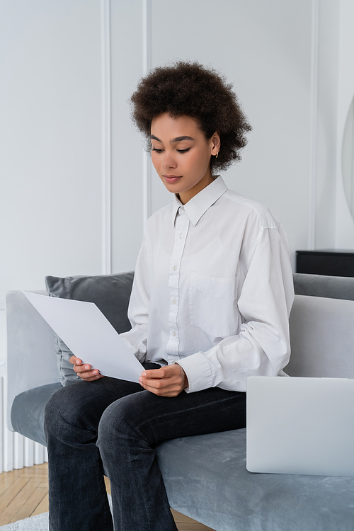 african american woman looking at blank document near laptop on velvet grey sofa