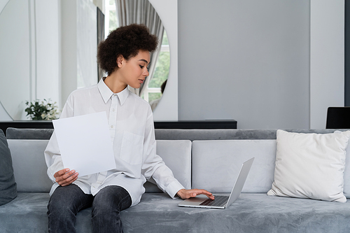 african american woman looking at blank document and using laptop on velvet grey sofa
