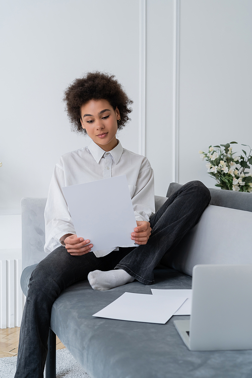 curly african american woman looking at blank document near laptop on velvet grey sofa