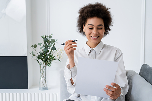 smiling african american woman holding pen and blank document while working from home