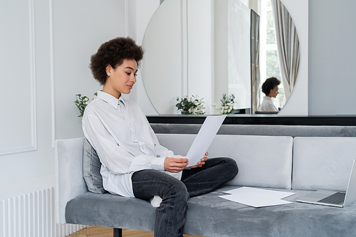 curly african american woman holding blank document near laptop on sofa while working from home