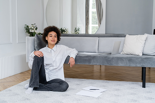 curly african american woman sitting near laptop and documents on carpet