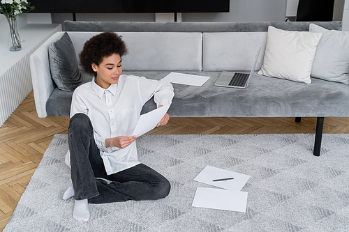 african american woman holding blank document while sitting near laptop on velvet sofa