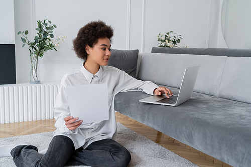 curly african american freelancer holding paper and using laptop on grey velvet sofa