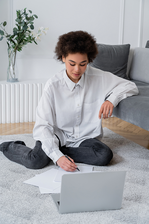 african american woman writing on paper while sitting on carpet near laptop