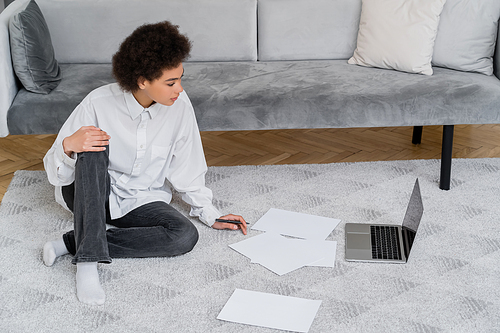 side view of african american woman looking at laptop while sitting on carpet near documents
