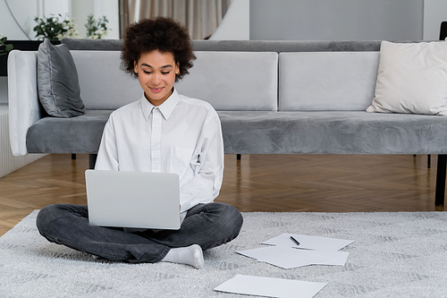 african american woman smiling while using laptop near documents on carpet
