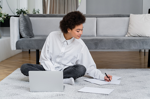 african american woman smiling while writing on paper and sitting with laptop on carpet