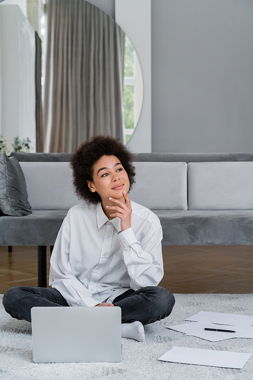 dreamy african american woman sitting near laptop and documents on carpet near grey velvet sofa