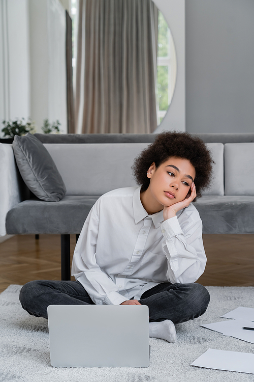 bored african american woman sitting near laptop and documents on carpet near grey velvet sofa