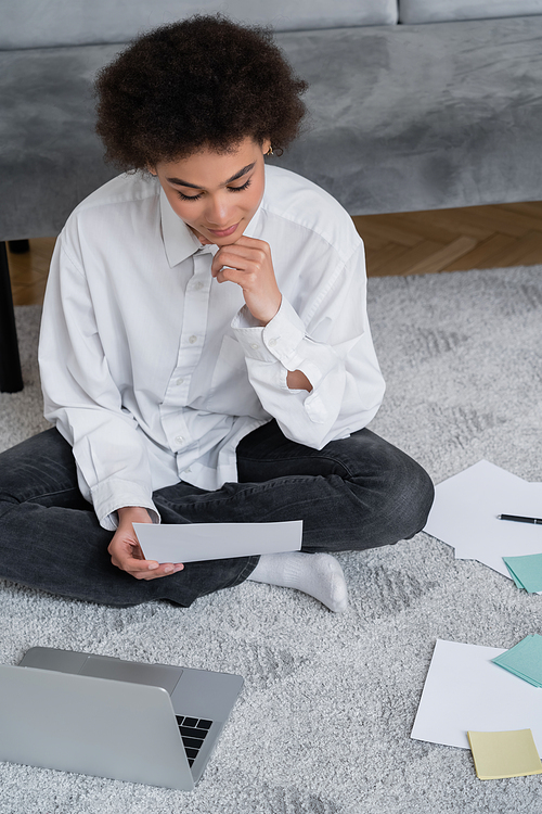 high angle view of curly african american woman looking at document and sitting on carpet near laptop