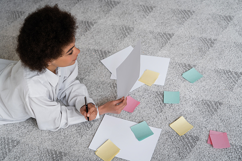 top view of curly african american woman holding document near blank papers on carpet