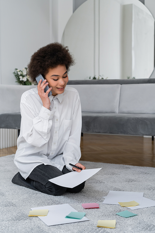 african american woman talking on smartphone while looking at document near blank papers on carpet