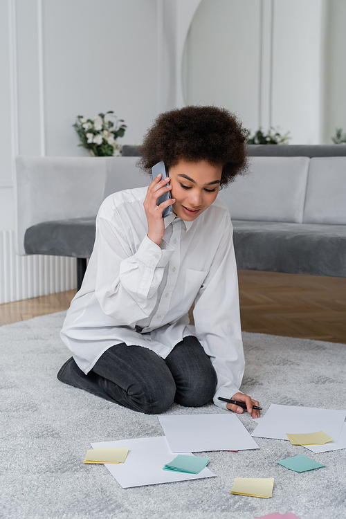 curly african american woman talking on smartphone while looking at document near blank papers on carpet