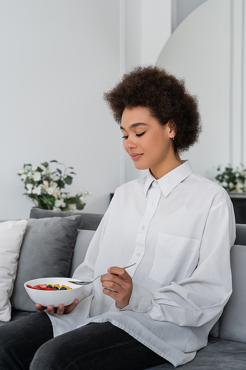 curly african american woman holding bowl with corn flakes and berries while having breakfast at home