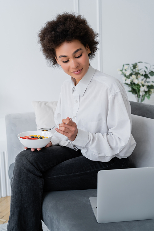 pleased african american woman holding bowl with berries while looking at laptop on velvet sofa