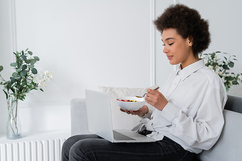 african american woman holding bowl with berries while watching movie on laptop and sitting velvet sofa