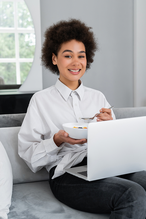 happy african american woman holding bowl with breakfast while looking at camera and sitting with laptop on velvet sofa