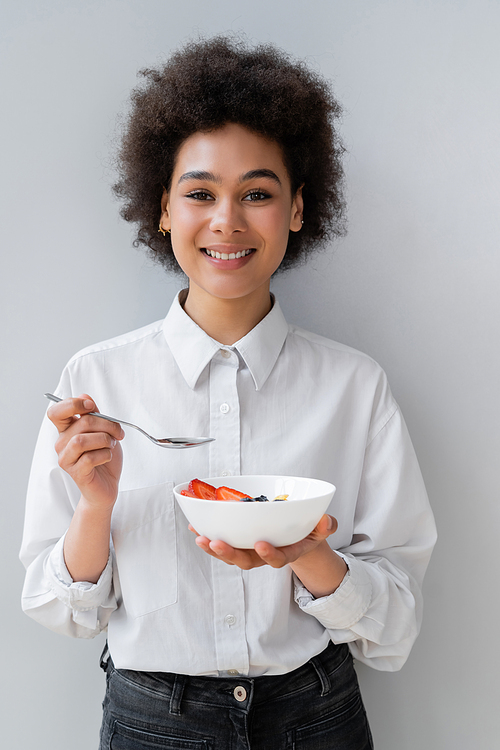 young and cheerful african american woman holding bowl with tasty berries on white
