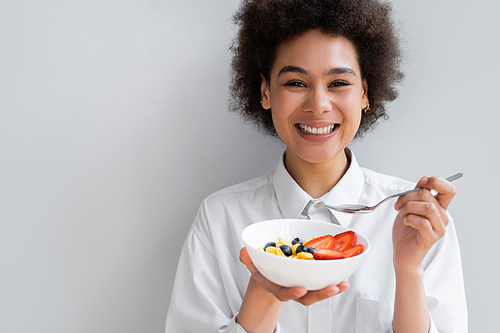happy african american woman holding bowl with corn flakes and berries on white