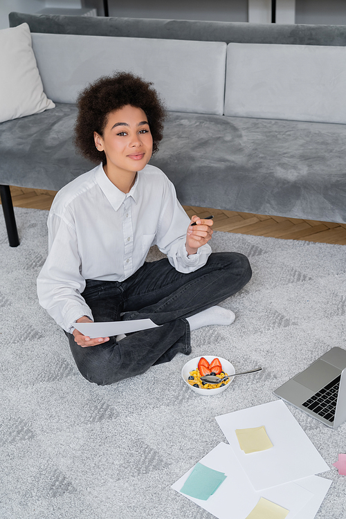 curly african american freelancer holding pen and paper while sitting on carpet near bowl with breakfast and laptop