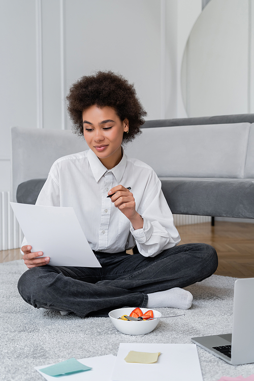 young african american freelancer holding pen and paper while sitting on carpet near bowl with breakfast and laptop