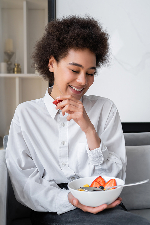 cheerful african american woman holding bowl with corn flakes and fresh strawberry