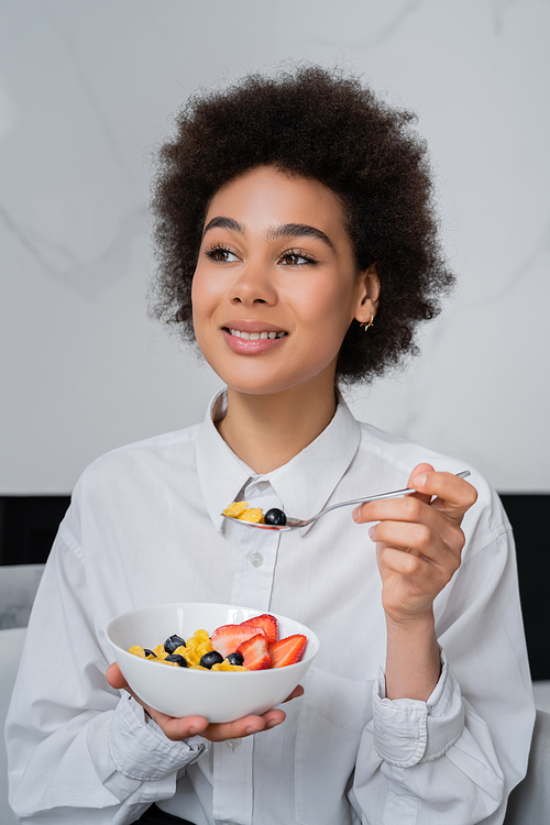 happy african american woman eating corn flakes with fresh berries at home