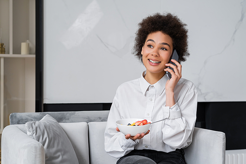 happy african american woman holding bowl with breakfast and talking on smartphone