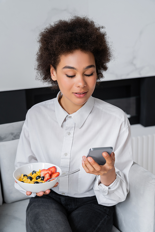 curly african american woman holding bowl with breakfast and smartphone