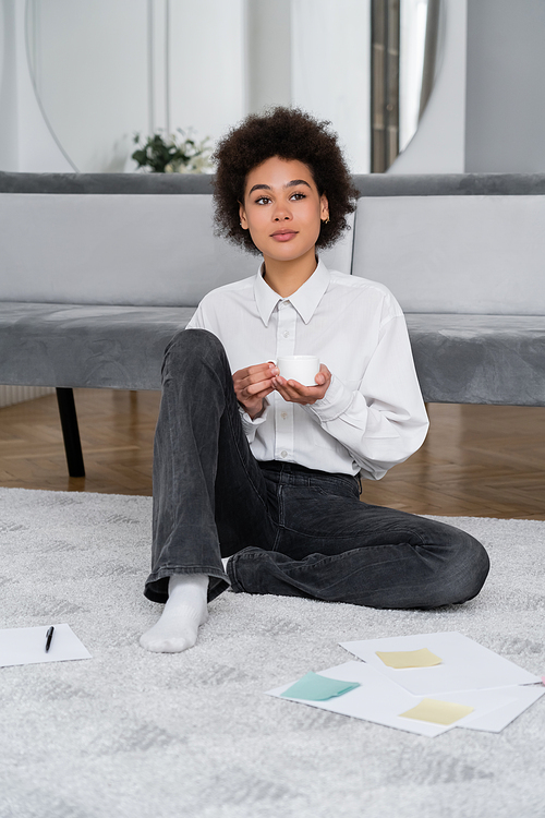 curly african american woman holding cup of coffee and sitting near documents on carpet