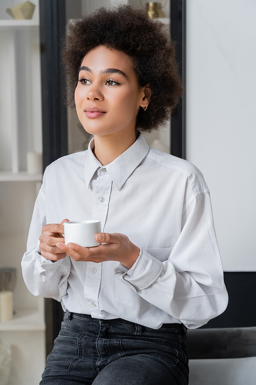 curly african american woman in white shirt with collar holding cup of coffee at home