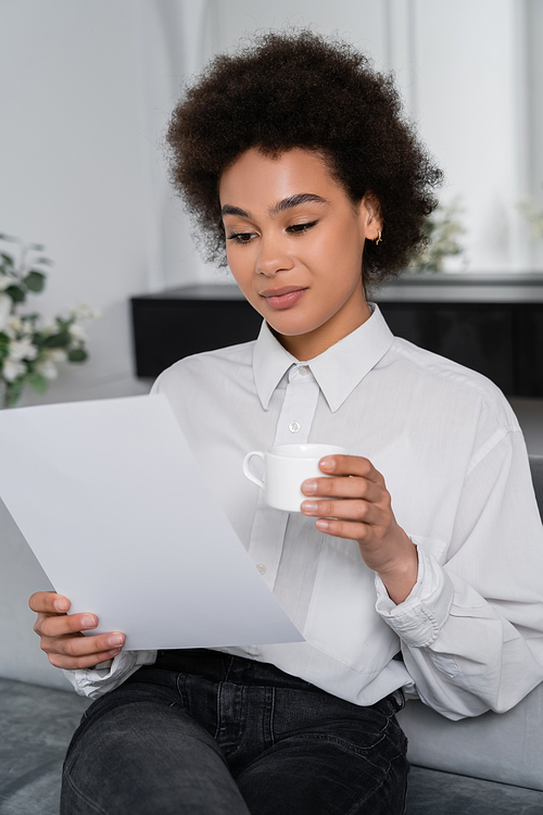 curly african american woman holding cup of coffee while looking at blank paper