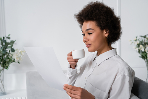 curly african american woman holding cup of coffee while looking at blank document