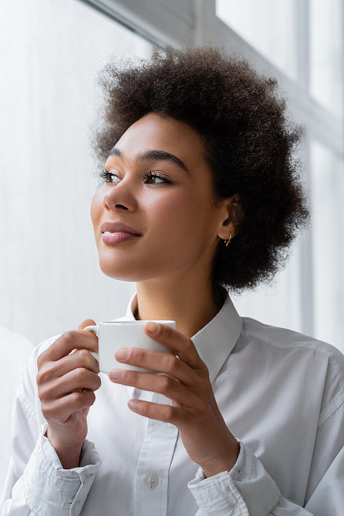 curly and smiling african american woman holding cup of coffee