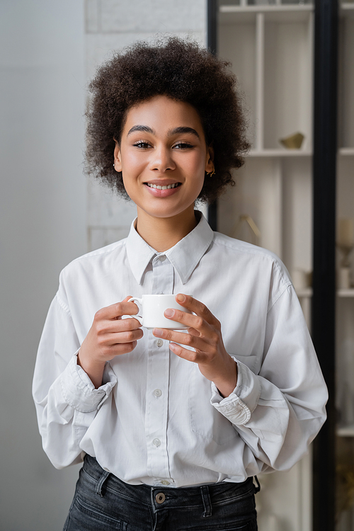 joyful and curly african american woman holding cup of coffee and looking at camera