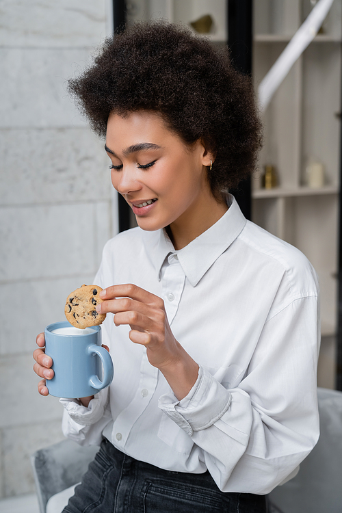 happy african american woman holding cup of milk and cookie with chocolate chips