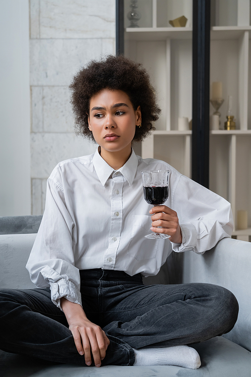 sad young african american woman holding glass of red wine while sitting on velvet sofa