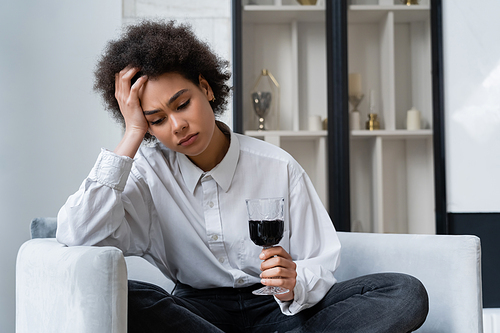 upset young african american woman holding glass of red wine while sitting on velvet sofa