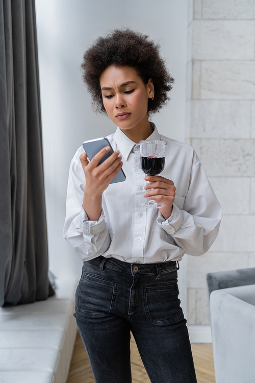 sad african american woman holding glass of red wine and looking at smartphone