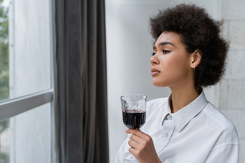 upset african american woman holding glass of red wine and looking at window