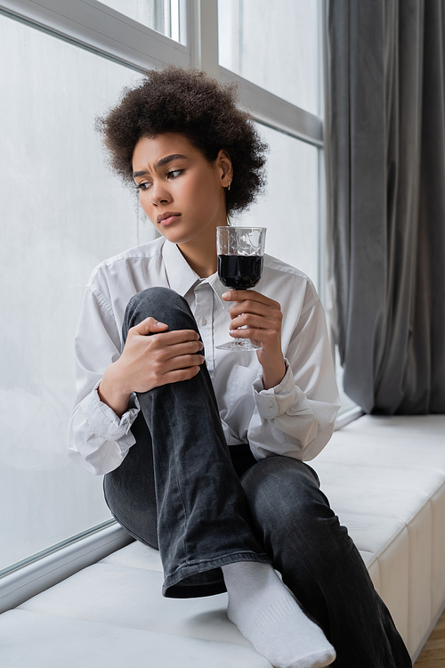 sad african american woman holding glass of red wine and looking at window while sitting at windowsill