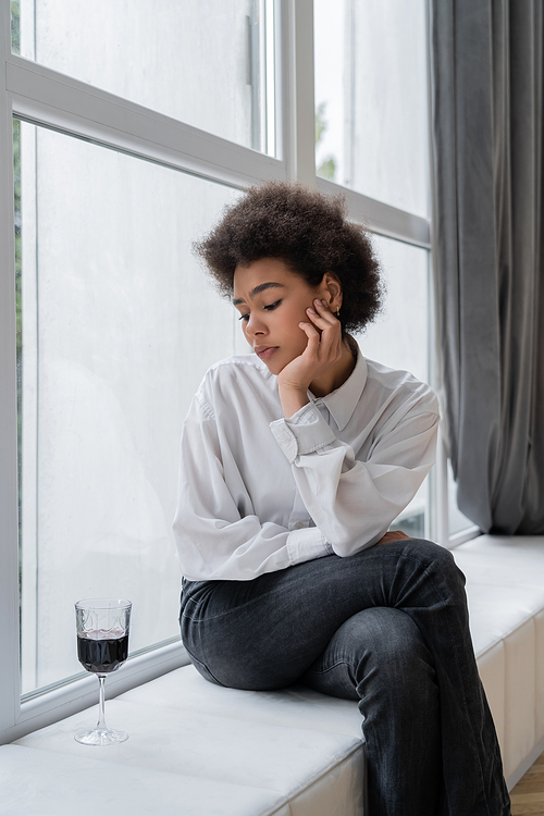 sad african american woman looking at glass of red wine while sitting on windowsill