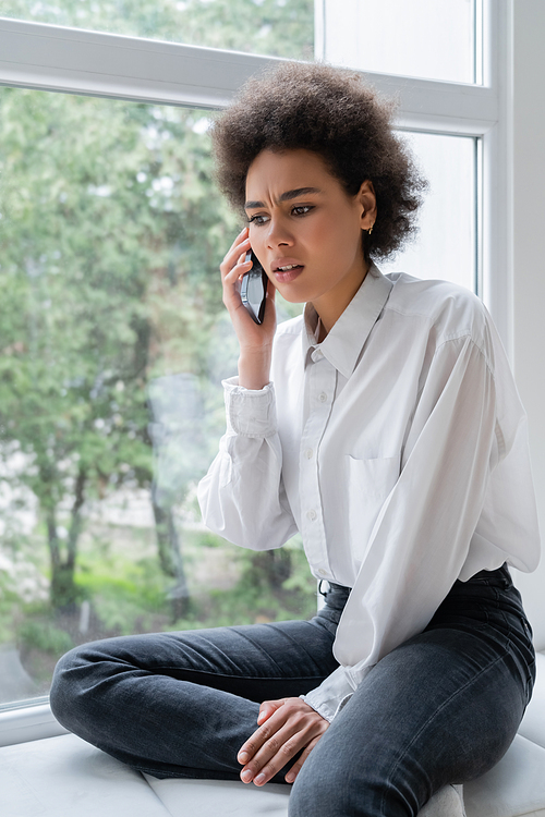displeased african american woman in white shirt talking on smartphone near window