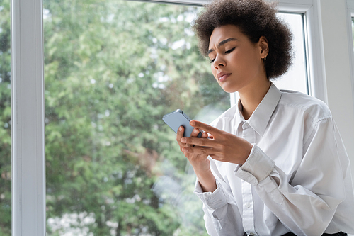 low angle view of upset african american woman in white shirt reading message on smartphone near window