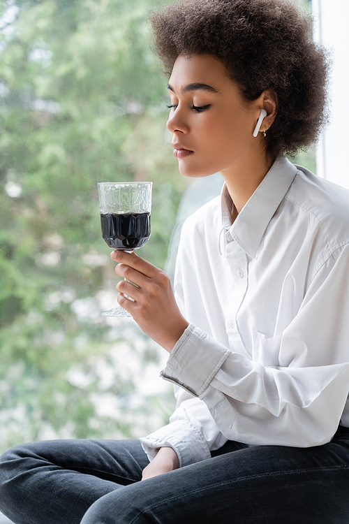 sad african american woman in white shirt and wireless earphone looking at glass of red wine