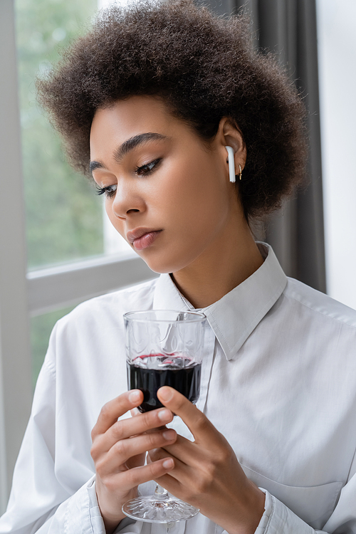 depressed african american woman in white shirt and wireless earphone holding glass of red wine