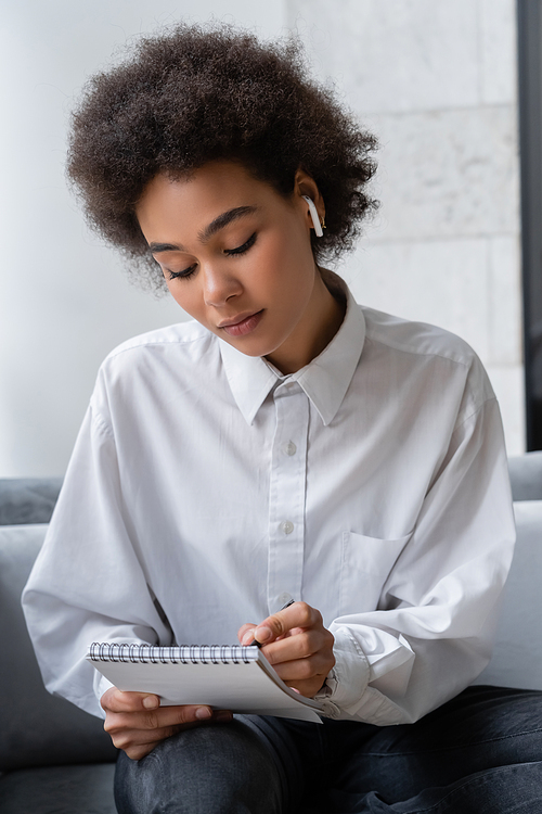 curly african american woman in white shirt and wireless earphone writing in notebook